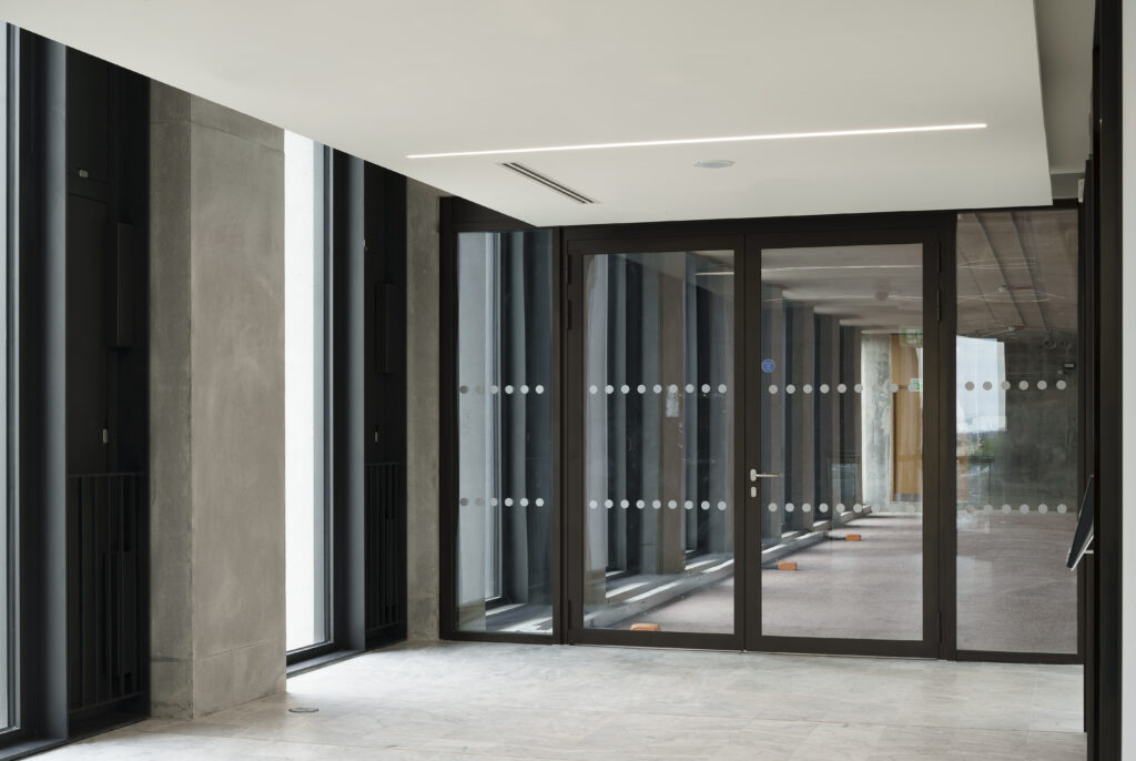 Inside a modern commercial building that hasn't been completed. Floors are tiled but walls are not painted. Looking at a full height double glass fire rated door with a same height glass screen either side. Through the doors we see a dip to a raw, unfinished floor ready for services to be installed.
