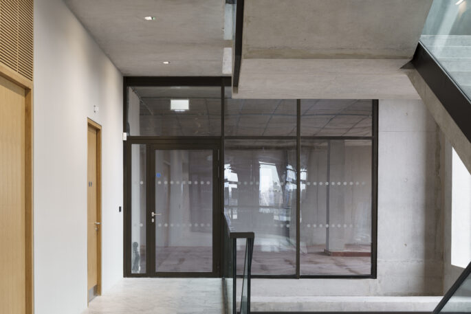 Interior of a commercial building looks still unfinished with plastered but not painted walls. Looking at a floor to ceiling height glass screen divided into three sections. On the far left the screen is replaced by a glazed door and slim screen. Two timber doors are visible on the left hand side.