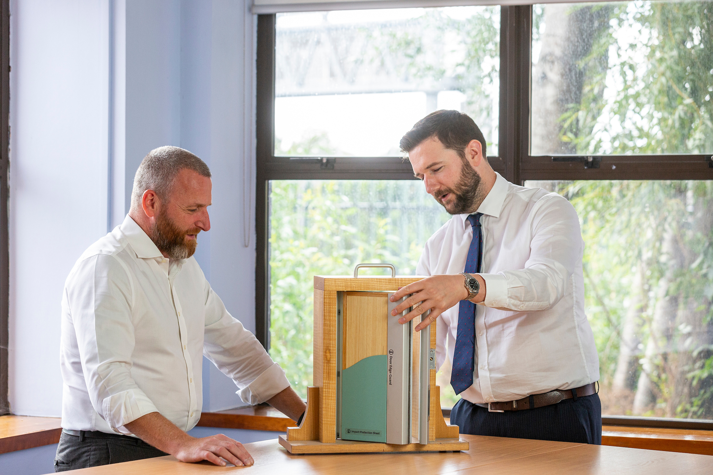 A picture of two men looking at a product sample on a table with windows in the background