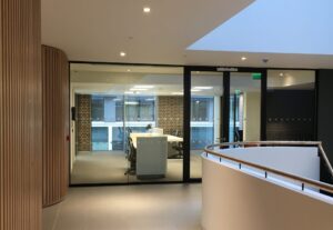 A modern building interior with a full height glazed screen and integrated double glazed door lead into an office area with big external windows overlooking another building. In the foreground of the image on the right is a curved half-height wall and stair-rail leading onto a staircase. On the left are two curved walls with modern timber cladding