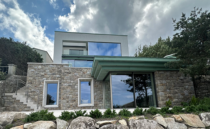 Looking up at the rear of a modern property spanning 3 storeys. On the lower ground level a glass sliding corner unit is visible. To the left are two tall single pane windows built into a decorative masonry wall. On the ground level above we see the top of large sliding glass windows which appear to lead onto a terrace area. A stairway up from the lower ground level patio is visible on the left. On the top floor we see a large glass window and a covered private balcony area. The property roof is not visible