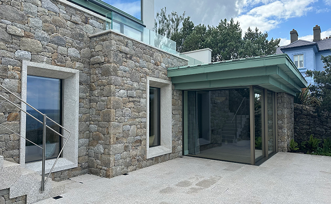 A view of the lower level of a house with a corner sliding window as the main focal point. The windows are in closed formation. To the left is a masonry wall with two tall single pane windows that sit in a picture frame type architrave in a lighter grey shade than the surrounding masonry