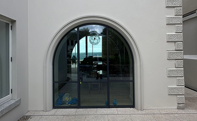 A view of an arched glass door with slim steel frame and two side screens built into a decorative plaster frame on the outside of a house.