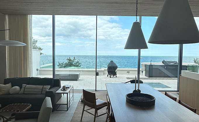 A view from inside a modern property looking out at a vast 3 metre tall sliding glass window system that slides back onto a fixed side screen opening up a large outdoor terrace area overlooking the sea.