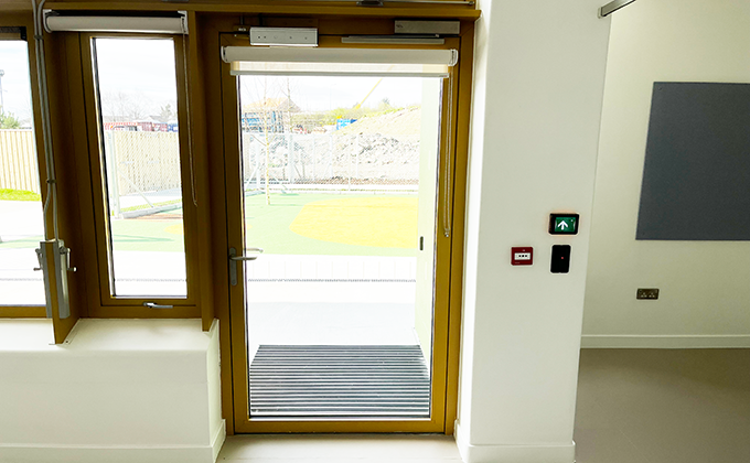 A straight on view from inside of a single glazed door on the outside of a special needs school which leads out into an outdoor play area. There is a section of wall to the right of the door with a wall reader and emergency release button to control the function of the door. An electric locking device is visible at the top of the door on the frame
