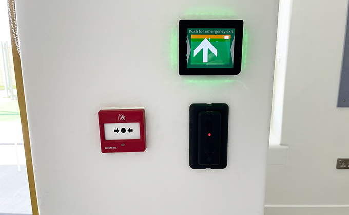 A wall showing three devices. We see a close up of an access control wall reader, a fire alarm push button and an emergency exit push button lit up in green.