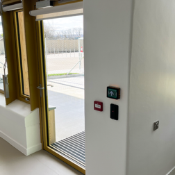 A view of a single external exit door leading out to an outdoor play area of a school. The door is glass and an electronic locking device is visible at the top and there is a wall reader for a fob or keycard on the wall next to the door.