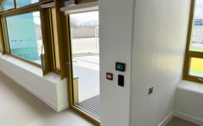 A view of a single external exit door leading out to an outdoor play area of a school. The door is glass and an electronic locking device is visible at the top and there is a wall reader for a fob or keycard on the wall next to the door.