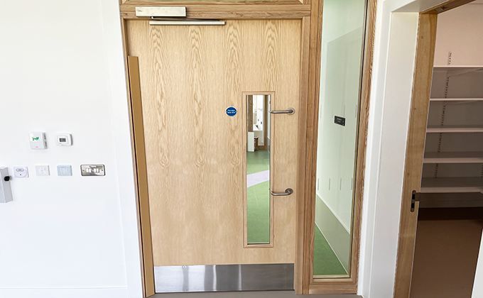 A view of a single wooden door with a slim glass panel close to one side. The door has a long hinge protector on it and two door handles, one at normal height and one at head height commonly seen in schools with special needs children. To one side of the door is a tall thin window showing the room behind the door.