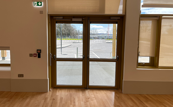 A view of a double glass door which leads out onto a school carpark or play area. The door has access control and emergency escape panic hardware on it including push bars.