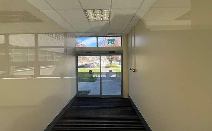 Looking out from a building corridor onto a single sliding automatic door with an adjacent glass screen and two narrow windows are visible overhead. The corridor is only very slightly wider than the door and screen combined.