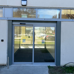 A view of a single automatic door with an adjacent glass screen on the outside of a building. There is a line of narrow windows above the door and behind the door we see a short hallway with another single automatic door and glass screen at the end of it