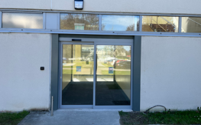 A view of a single automatic door with an adjacent glass screen on the outside of a building. There is a line of narrow windows above the door and behind the door we see a short hallway with another single automatic door and glass screen at the end of it