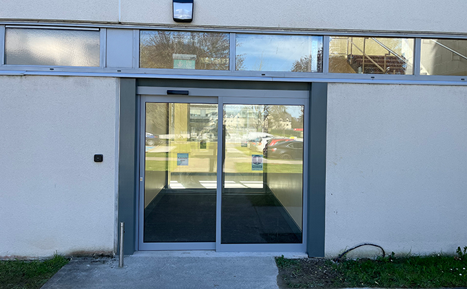 A view of a single automatic door with an adjacent glass screen on the outside of a building. There is a line of narrow windows above the door and behind the door we see a short hallway with another single automatic door and glass screen at the end of it